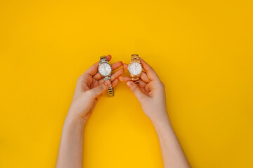 Female hands holding two wrist watches, golden and silver, on yellow background.