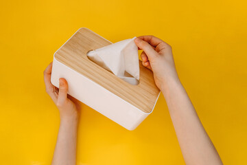 Female hands taking a paper tissue out of a wooden box, on yellow background.