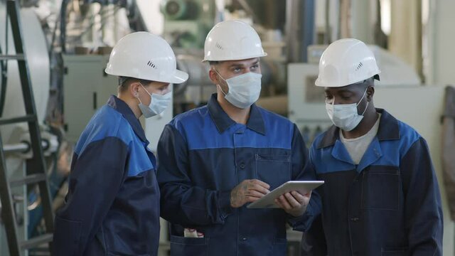  Slowmo Medium Shot Of Female And Male Plant Workers In Face Masks, Hard Hats And Uniforms Looking At Tablet And Having Conversation At Workplace