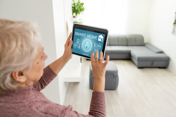 Senior woman at home standing at kitchen holding digital tablet controlling smart home system back...