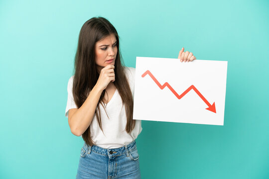 Young caucasian woman isolated on blue background holding a sign with a decreasing statistics arrow symbol and thinking