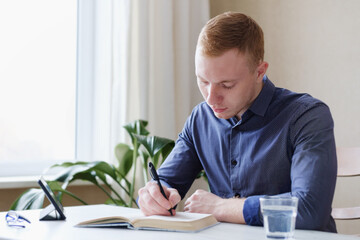 Concentrated businessman writing notes in notebook and watching webinar video course on tablet - Young man studying via video conference - Distance learning and online education concept
