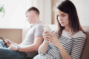 Disgruntled husband and wife or boyfriend and girlfriend are sitting on the couch and looking at the phone - Annoyed young people ignore each other and use smartphones - Bored young couple with gadget
