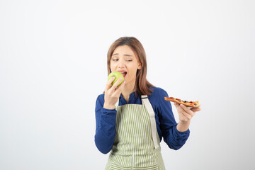 Portrait of woman in apron eating green apple while looking at pizza