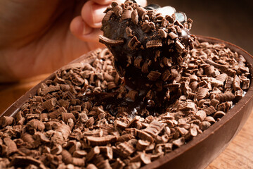 Close up of a hand sticking a metallic spoon in a stuffed chocolate easter egg with grated chocolate on the top on a wooden stand on a wooden table. 
