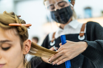 Obraz na płótnie Canvas Asian professional female stylist cutting Caucasian woman's hair in salon. The man wearing mask and face shield to prevent from coronavirus infection during pandemic. Beauty salon business concept.