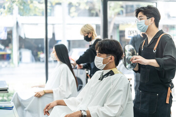 Professional male stylist cutting man's hair in salon. The man wearing mask and face shield to...