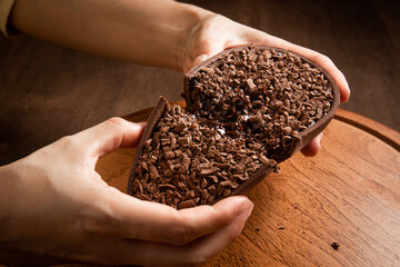 Woman hands crack a stuffed chocolate easter egg with grated chocolate on the top on a wooden stand on a wooden table.