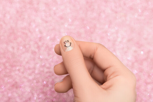 Female Hand With Pink Nail Design. Smiling Face Nail Art Decoration On A Thumb. Woman Hand On Glitter Pink Background.