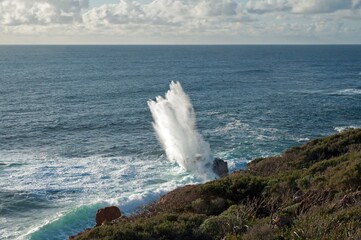 A high vertical plume of seawater exploding above a huge rock after a large wave has hit it.  
Seascape of a calm ocean with green vegetation in the foreground.