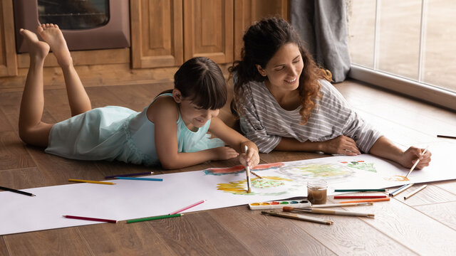 Overjoyed young Latino mother and small 8s daughter lying on floor at home have fun painting together with colors. Happy Hispanic mom or nanny and little girl child enjoy drawing. Hobby concept.