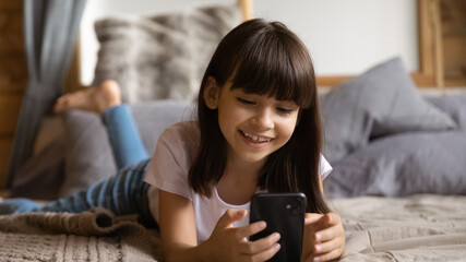 Obraz premium Smiling Latino 8s kid lying on bed using cellphone gadget have online webcam digital virtual conference. Happy Hispanic girl child hold smartphone speak talk on video call on modern device.