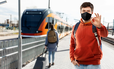 travel, tourism and pandemic concept - young man with backpack in face protective medical mask showing ok hand sign traveling by train over railway station in city of tallinn, estonia on background