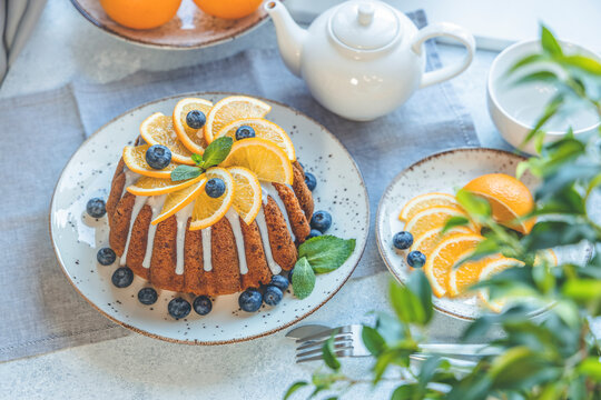 Orange bundt cake with blueberry surrounded fruits, plant and cutlery on light table near window. Family breakfast concept