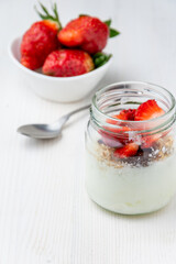 Top view of jar with yogurt, strawberries and muesli, spoon and bowl with strawberries, selective focus, on white wooden table, vertical