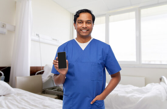 medicine, healthcare and technology concept - happy smiling indian doctor or male nurse in blue uniform with smartphone over hospital ward background
