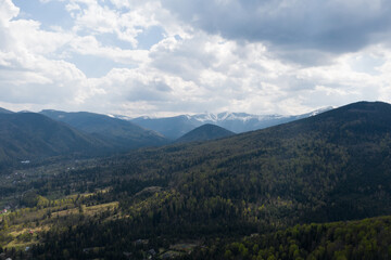 Fototapeta premium Summer mountain landscape, beautiful scenery of Carpathian mountains in Western Ukraine. High mountain peaks covered with snow, cloudy sky, green forests and a small village at the foot of the hill.