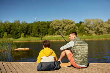 family, generation, summer holidays and people concept - happy smiling father and son with fishing rods on river berth