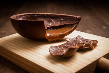 Cracked chocolate easter egg with flower of salt, stuffed with dulce de leche (dripping) on a wooden stand on a wooden table.