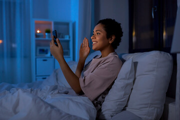technology, internet, communication and people concept - happy smiling young african american woman with smartphone having video call lying in bed at home at night