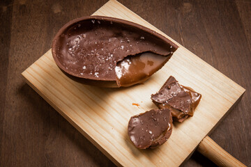 Cracked chocolate easter egg with flower of salt, stuffed with dulce de leche (dripping) on a wooden stand on a wooden table.