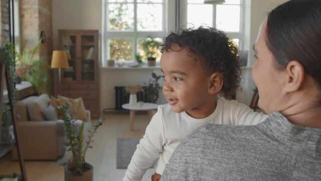 Slowmo shot of cheerful mother holding adorable black toddler boy and talking to him while he is looking out window with curiosity