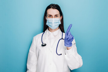Serious professional woman doctor in protective face mask and gloves warning people, shaking finger in prohibition restriction, isolated on blue wall. Medical workers, pandemic coronavirus concept