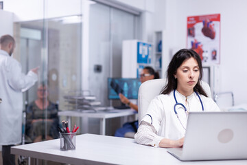 Serious young woman doctor physician wearing white coat using laptop computer writing notes at workplace. Female professional medic consulting patient in online chat sit at desk in hospital.