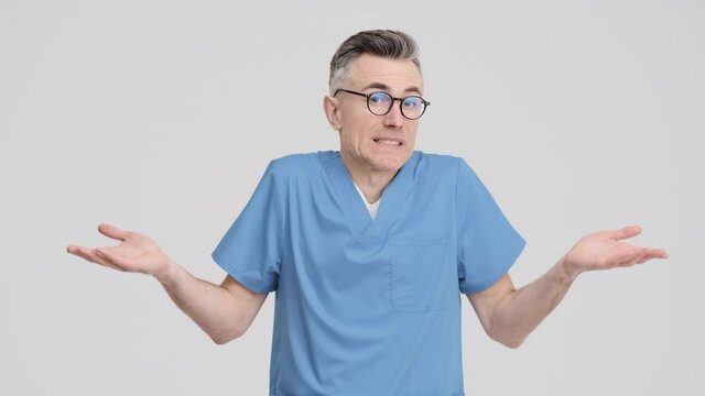 A confused doctor man wearing uniform is shrugging shoulders standing isolated over grey background in the studio