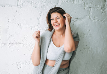 Beautiful happy emotional brunette young woman plus size body positive in comfortable sport wear near white brick wall