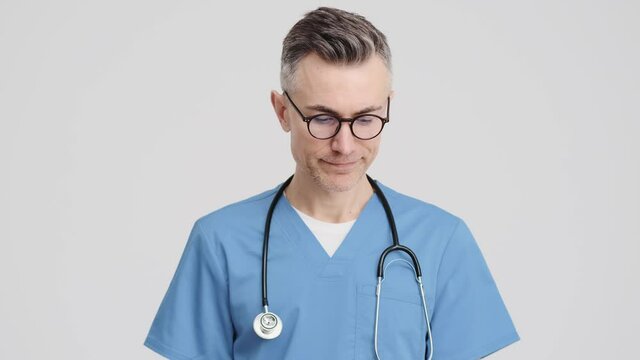 A displeased senior doctor man wearing medical uniform is shaking his hand negatively standing isolated over grey background in the studio