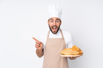 Male baker holding a table with several breads isolated on white background pointing finger to the side