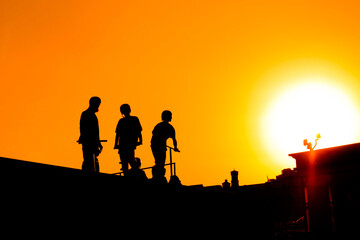 Unrecognizable group of teenage boy silhouettes with scooters standing together against orange sunset sky at skatepark. Sport, extreme, freestyle, outdoor activity concept