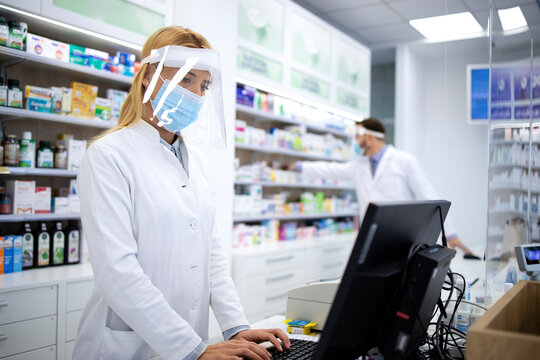 Female blonde pharmacist cashier wearing face shield and white coat selling vitamins in pharmacy store during corona virus pandemic.