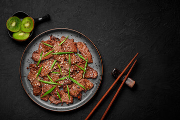 Beef Bulgogi on black plate on dark slate table top. Korean cuisine meat dish with sauce and kiwi. Traditional asian food. Top view. Copy space