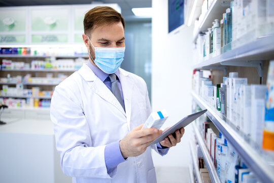 Pharmacist in protection mask holding tablet computer and checking medicines in pharmacy shop.