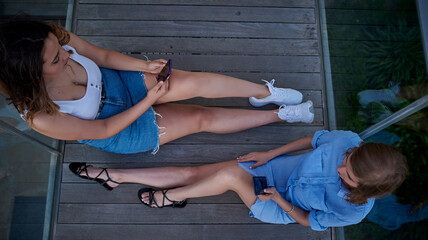 Aerial overhead shot of two young women with their smart phones in hand. They are sitting on a wooden floor.