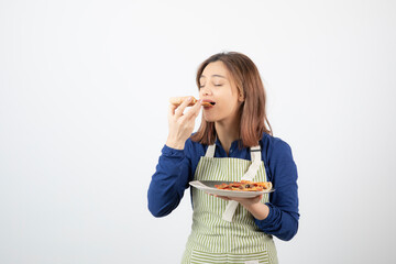 Shot of female cook in apron eating slice of pizza on white background