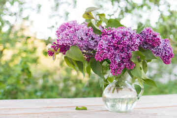 Fresh lilacs are arranged in a glass vase and placed on an outdoor table.