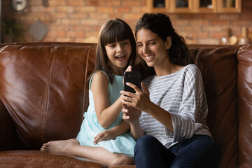 Happy little girl child relax on sofa at home with young Latino mother talk on video call on cellphone. Smiling Hispanic mom with small daughter have fun engaged in webcam online digital conversation.