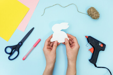 DIY easter decoration. Step-by-step instruction: how to create a garland made of bunnies. Handmade Easter craft. Hands holding paper bunny template around necessary materials. Step 1.