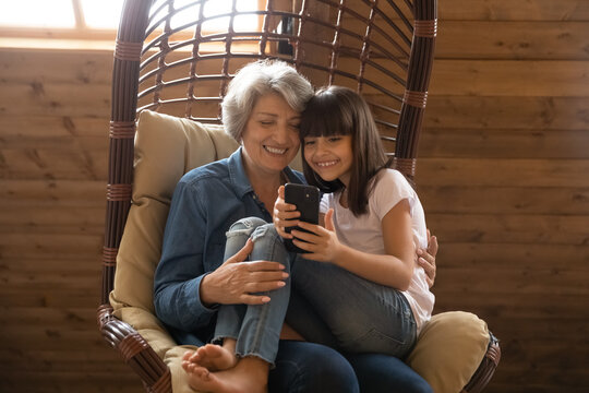 Caring smiling mature Hispanic grandmother and small teen granddaughter relax at home watch video on cellphone. Happy elderly Latino grandparent and 9s grandchild have fun using smartphone.