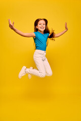 Fototapeta na wymiar Positive girl wearing a blue t-shirt and white jeans, jumping with raised hands, on yellow background