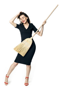 Beautiful brunette woman in black dress and red shoes playing a broomstick like a guitar, like a housewife isolated on a white background