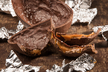 Cracked stuffed chocolate easter egg with salt of flower and filled with dulce de leche on a wooden table. Ripped aluminium paper around it.