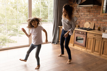 Happy young Latino mother and excited little 8s daughter dance together in modern renovated kitchen together. Overjoyed Hispanic mom or nanny have fun with small teen girl child at home. Rent concept.