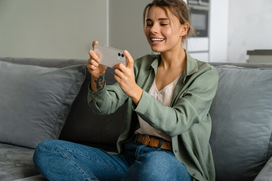 Happy Woman Playing Online Game On Mobile Phone While Sitting On Sofa