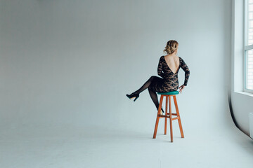 A young woman in a beautiful black guipure dress with an open back sits on a bar stool with her back to the viewer in an empty gray room