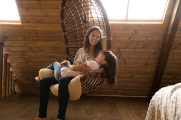 Caring young Hispanic mother and little 8s daughter sit relax in swing cozy rattan chair at home...