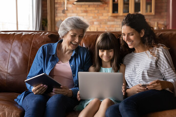 Happy three generations of Latino women sit relax on couch at home using modern electronic gadgets...